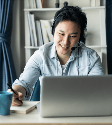 Man wearing headset smiling at a laptop screen, holding a pen to a notepad