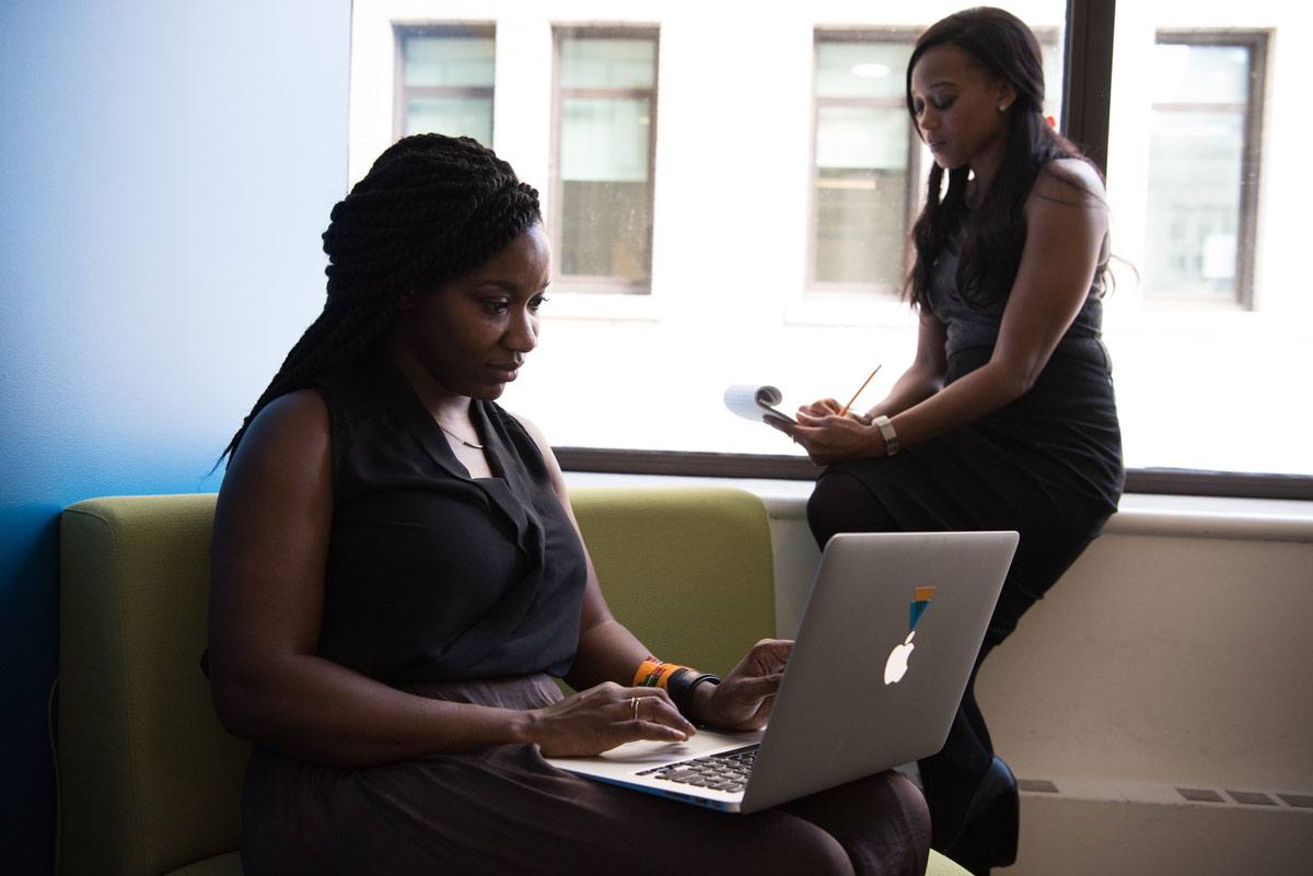 A woman working on a laptop, and another woman writing on a notepad in the background